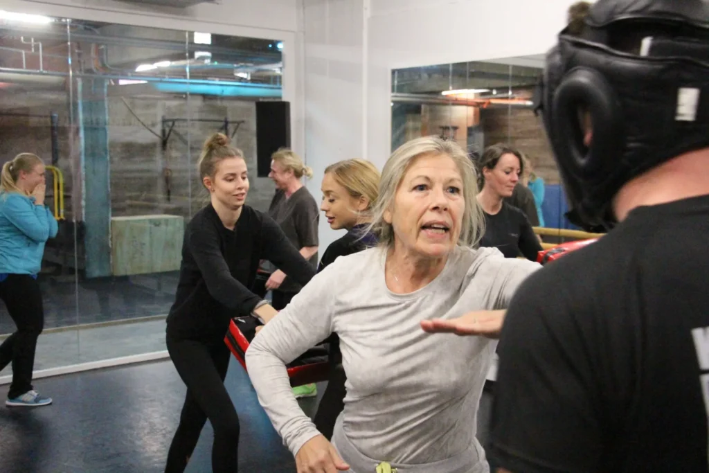 Dame mit grauen Haaren, die engagiert einem Mann mit Trainingshelm auf die Stirn bietet, im Hintergrund weitere Frauen, die auf Schlagpolster treten